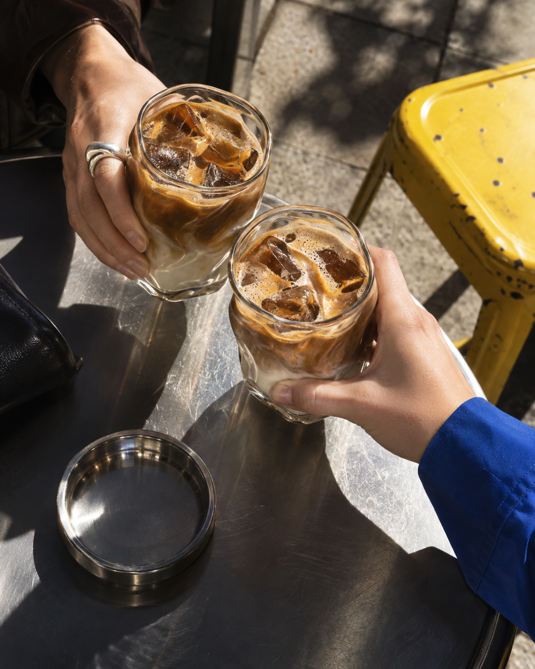 Dos personas brindan con café helado sobre una mesa metálica, junto a un taburete amarillo.